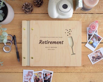 May include: A rectangular light-colored wooden photo album with a metal spine and gold-tone fasteners. The cover features the text "CONGRATULATIONS ON YOUR RETIREMENT", "KELLY BROWN", and "2003-2024", alongside a laser-cut champagne flute graphic.