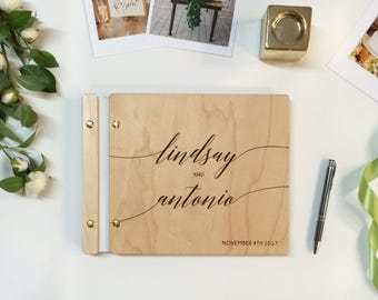 Peut inclure: Un album photo en bois clair avec une reliure en métal doré. La couverture présente les noms gravés "lindsay" et "antonio" en écriture cursive, avec le texte "AND" et la date "NOVEMBER 4TH 2017" inscrits en dessous.