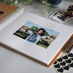 A graduation guest book lies open on a table with a 4x6 photograph of the graduate in their cap and gown. Photo holders lie next to the guest book for posting photo booth strips, polaroid pictures, and scrapbooking material.