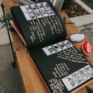 An 8.5x11 inch guest book lies open on a wooden desk. Black cardstock pages with handwritten messages and photo booth strips are posted on the pages for the bride and groom to cherish as a keepsake after their wedding day.