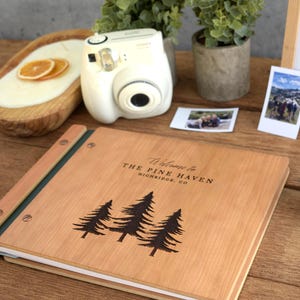 May include: A square wooden photo album with a light brown finish, featuring a laser-engraved design of three pine trees and the text "Welcome to THE PINE HAVEN HIGHRIDGE, CO" on the front cover, secured with metal binding screws.