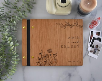 May include: A rectangular wooden photo album with a light brown grain, featuring a black spine secured by four gold-toned metal rivets. The cover is engraved with a floral design, the text "AMIN and KELSEY", and the date "08/26/23" in black ink.