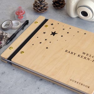 May include: A wooden guest book with a black spine and gold accents. The cover features a star design and the text "Welcome Baby Kern-Hill 2025" and "Guestbook".