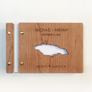 May include: A rectangular wooden photo album with a light brown grain, featuring a cutout of the island of Jamaica in the centre. The cover is engraved with the text MICHAEL + MIRYAH, NOVEMBER 12, 2016, and NEGRIL HEART JAMAICA, held by brass screws.