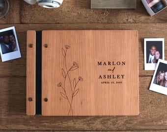 May include: A rectangular wooden photo album with a light brown grain, featuring a black spine and metal binding screws. The cover is engraved with a line-art floral design and the text MARLON and ASHLEY APRIL 13, 2023 in black ink.