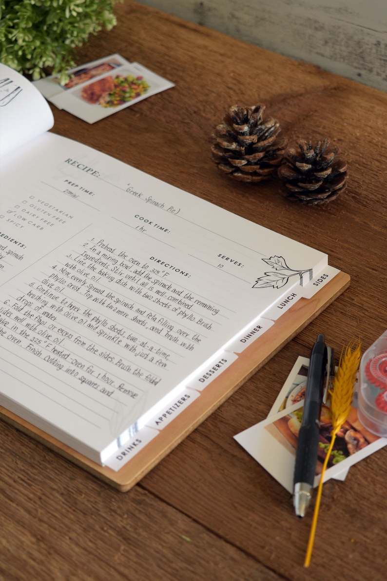 May include: A close-up of a recipe book with a wooden spine and tabs labeled with different meal categories. The recipe is for Greek Spinach Pie and includes a list of ingredients, directions, and serving size. The book is open on a wooden table with a pen and other items.