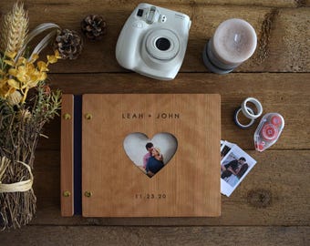 May include: A rectangular wooden photo album with a heart-shaped cutout in the center displaying a photograph of two people. The cover is engraved with the text LEAH + JOHN and the date 11.23.20, featuring gold-toned metal binding screws.