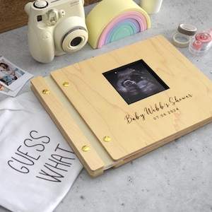 May include: A light-coloured wooden photo album with gold-toned metal binding screws, featuring a square cutout displaying a black and white ultrasound image. The cover is engraved with the text Baby Webb's Shower and the date 07.06.2024.