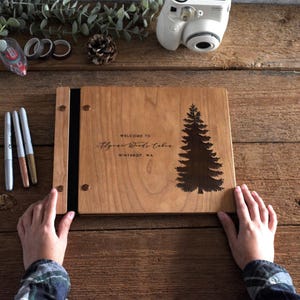 May include: A rectangular wooden guest book with a light brown finish, featuring a dark engraved pine tree graphic on the right side and the text "WELCOME TO Alpine Woods Cabin WINTHROP, WA" centered on the front cover, secured with metal fasteners.