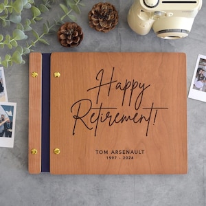 May include: A rectangular wooden photo album with a dark blue spine and gold-colored metal binding screws. The front cover features the black engraved text "Happy Retirement!" followed by "TOM ARSENAULT" and the years "1997 - 2024" below it.