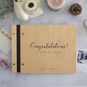 Pode incluir: Um álbum de fotos retangular de madeira com acabamento em grão de madeira clara. A capa apresenta o texto "Congratulations! ON YOUR RETIREMENT" e o nome "CADY WILSON" em letras castanho-escuras. É fixado com parafusos de metal dourado.