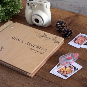 May include: A wooden recipe book with the text "Mom's Favorite Recipes" engraved on the cover. The book is open and there are photos of food on the table next to it.