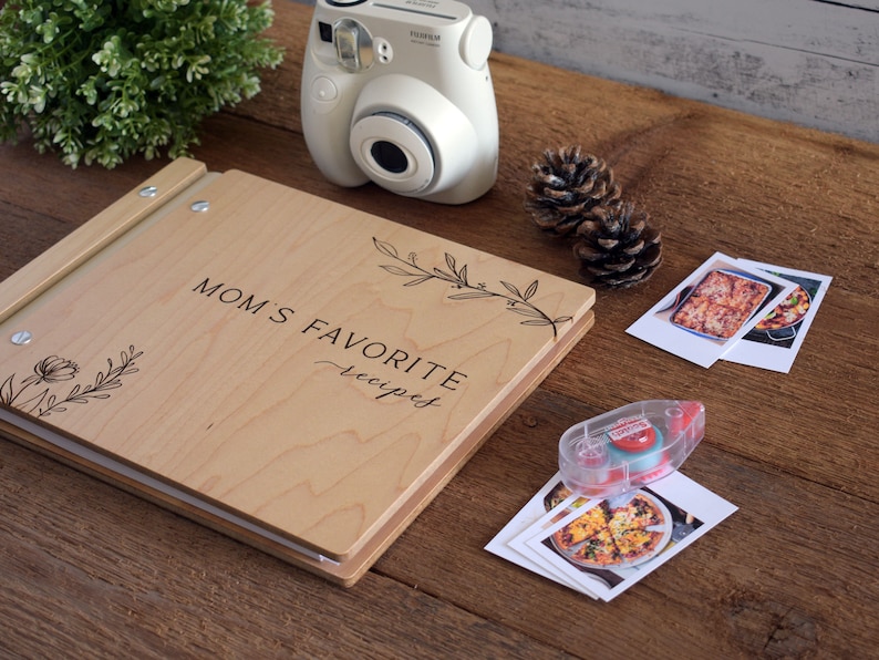 May include: A wooden recipe book with the text "Mom's Favorite Recipes" engraved on the cover. The book is open and there are photos of food on the table next to it.