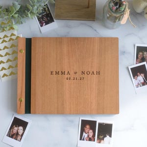 Könnte beinhalten: Ein rechteckiges Fotoalbum aus Holz mit einem dunkelgrünen Buchrücken und goldfarbenen Metallschrauben zur Bindung. Auf dem Vorderdeckel ist der Text EMMA & NOAH sowie das Datum 05.21.27 in einer zentrierten, dunklen serifenlosen Schrift eingraviert.