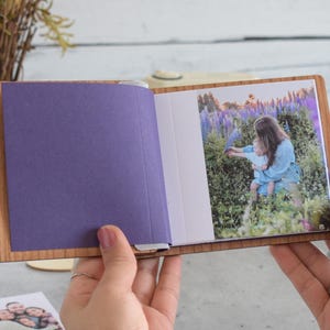 May include: A small, open photo album with light brown wooden covers. The left side features a solid purple page, while the right side displays a color photograph of a person holding a child in a field of purple lupine flowers and green foliage.