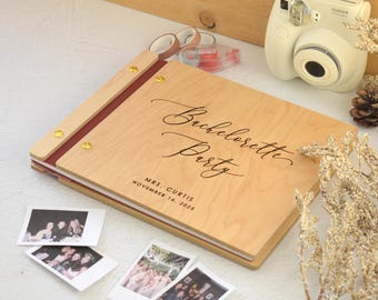 Op de afbeelding: Een fotoalbum van licht hout met een donkerrode rug en gouden metalen schroeven. De kaft bevat de gegraveerde tekst Bachelorette Party, Mrs. Curtis en November 16, 2023. Het album wordt getoond met verschillende kleine vierkante instant foto's.