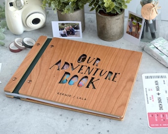May include: A rectangular wooden photo album with a dark green spine and metal binding screws. The front cover features laser-cut text reading OUR ADVENTURE BOOK and the names KERMIE / LALA. The wood grain is visible, and the cover has a cutout design.