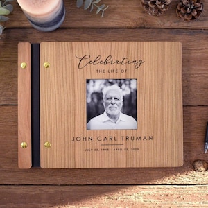 May include: A wooden photo album with a light brown grain finish, featuring a central black and white portrait of a person. The cover text reads Celebrating the life of John Carl Truman, July 23, 1945 - April 03, 2023, with gold metal fasteners.