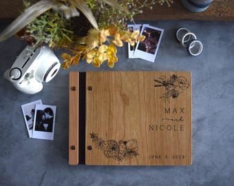 May include: A rectangular wooden photo album with a light brown grain, featuring a dark spine and metal binding screws. The cover is engraved with floral line art, the names MAX and NICOLE, and the date JUNE 3 2023 in a clean, black font.
