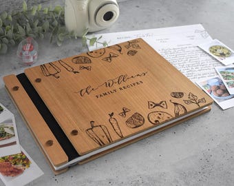Puede incluir: Un libro de recetas cuadrado de madera con acabado marrón claro, que presenta el texto grabado "the Williams FAMILY RECIPES" rodeado de ilustraciones de verduras y pasta. La cubierta está asegurada con cierres metálicos y encuadernación negra.