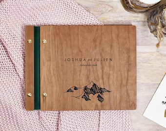 May include: A rectangular wooden photo album with a dark green spine and gold-toned metal binding screws. The front cover features the engraved text JOSHUA and JULIEN Adventure book above a black line drawing of a mountain range.