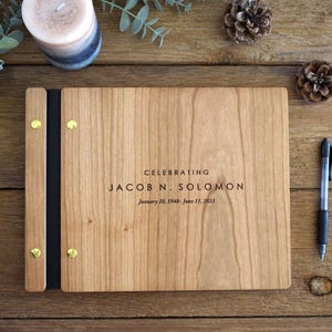 May include: A rectangular wooden memorial book with a light brown grain, featuring a black spine secured by four gold-toned metal screws. The cover is engraved with the text: CELEBRATING JACOB N. SOLOMON January 30, 1948 - June 11, 2023.