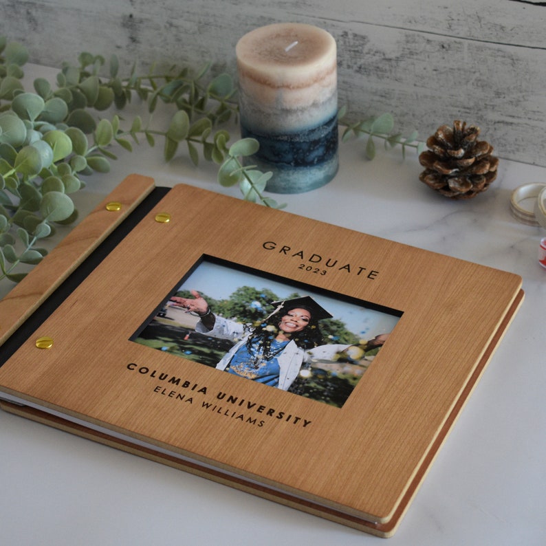 A graduation photo album lies on a table with polaroid photos of the graduate, washi tape, and double sided tape. This book serves as a graduation party decoration for guests to sign and post polaroids inside. It also makes the perfect grad gift.
