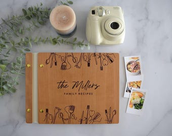 May include: A wooden recipe binder with gold-tone metal fasteners, featuring the engraved text "the Millers FAMILY RECIPES" and black line art illustrations of kitchen utensils along the top and bottom edges of the front cover.