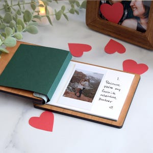 a small photo book with a handwritten note, a green paper, and a wooden frame, all placed on a marble surface.