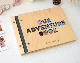 May include: A light-colored wooden photo album with a dark green spine and metal fasteners. The cover features laser-cut lettering that reads OUR ADVENTURE BOOK, revealing a landscape photo underneath, with the names LUCAS and SHAYLA printed below.