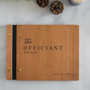May include: A rectangular wooden binder with a light brown wood grain finish, featuring a black spine secured by four gold-toned metal screws. The cover is engraved with the text "the OFFICIANT", "Lanie Dornie", and "MICHAEL + KRYSTAN 12.09.2023".