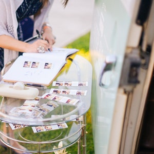 An 8.5x11 inch guest book lies open on a glass table as a female wedding guest signs their name and issues handwritten messages underneath photobooth strips in order to congratulate the newlyweds.