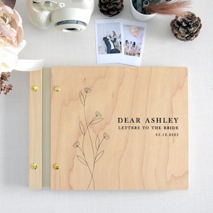Puede incluir: Un álbum de recortes rectangular de madera clara con una tira de encuadernación de metal dorado y cuatro tornillos dorados. La cubierta presenta una ilustración floral de líneas negras y el texto: DEAR ASHLEY LETTERS TO THE BRIDE 05.12.2025.