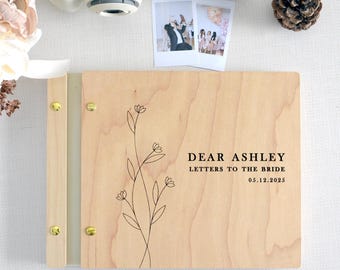May include: A rectangular light-colored wooden scrapbook with a gold-tone metal binding strip and four gold-tone screws. The cover features a black line-art floral illustration and the text: DEAR ASHLEY LETTERS TO THE BRIDE 05.12.2025.
