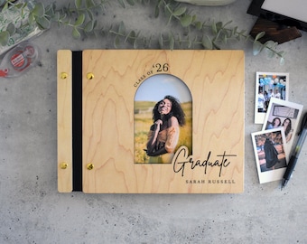 May include: A light-colored wooden photo album with a vertical arch-shaped cutout displaying a portrait of a person. The cover features the text "CLASS OF '26", "Graduate", and "SARAH RUSSELL" in black, with gold-toned metal binding screws.