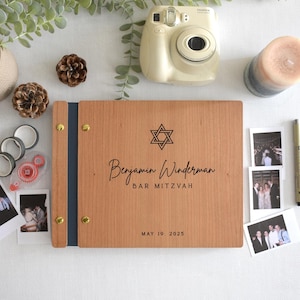 Puede incluir: Un álbum de fotos rectangular de madera con un lomo azul oscuro y tornillos de encuadernación de metal dorado. La cubierta presenta una estrella de David negra, el nombre Benjamin Winderman, el texto BAR MITZVAH y la fecha MAY 19, 2025, grabados.