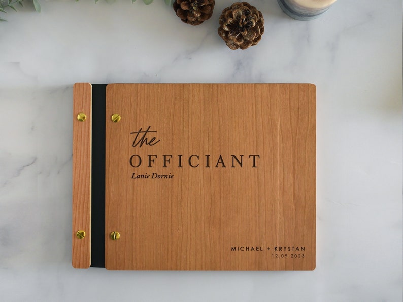 May include: A wooden album with a black leather spine and gold accents. The front cover is engraved with the text "the officiant Lanie Dornie" and "MICHAEL + KRYSTAN 12.09.2023".