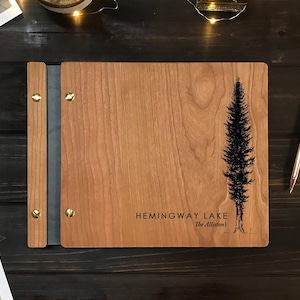 May include: A rectangular wooden photo album with a light brown wood grain finish. The cover features a black engraved pine tree graphic and the text "HEMINGWAY LAKE The Alliston's". It is secured with two gold-toned metal binding screws.