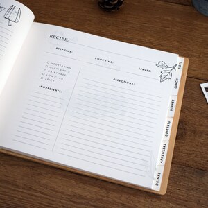 May include: A white recipe book with a wooden spine and tabs for different meal categories, including drinks, appetizers, desserts, dinner, lunch, and sides. The book has lines for writing down recipes and includes checkboxes for dietary restrictions like vegetarian, gluten-free, dairy-free, low carb, and spicy.