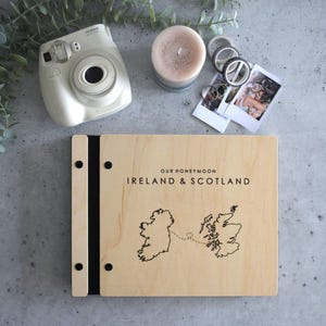 May include: A light-colored wooden photo album with black binding screws and a black spine. The cover features the text "OUR HONEYMOON IRELAND & SCOTLAND" above a black line-art map of Ireland and Scotland connected by a dashed line with a heart.
