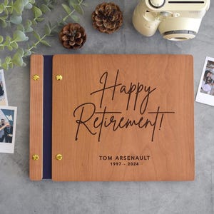 May include: A rectangular wooden photo album with a dark blue spine and gold-colored metal binding screws. The front cover features the black engraved text Happy Retirement! followed by the name TOM ARSENAULT and the years 1997 - 2024.