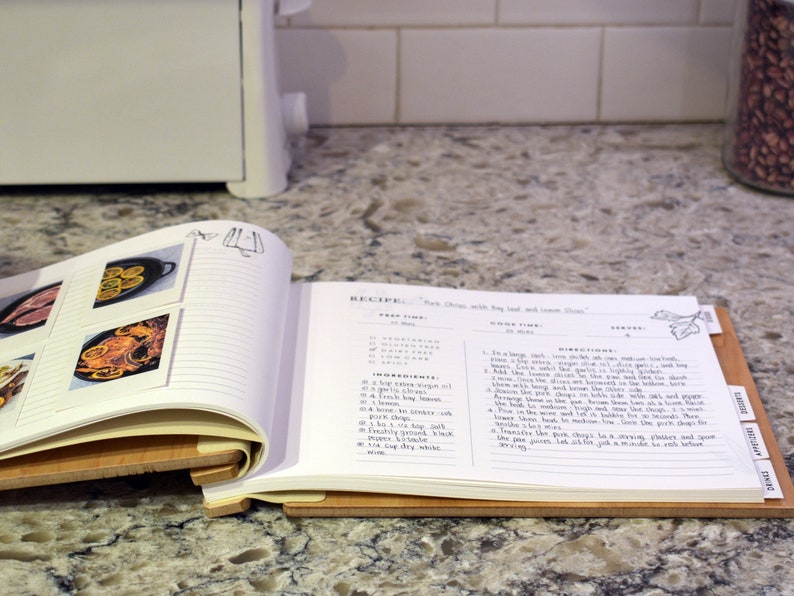 May include: Open recipe book with a wooden stand, showing a recipe for "Pork Chops with Bay leaf and Lemon Thyme". The recipe includes ingredients, prep time, cook time, and directions. The book has lined pages and photos of food.