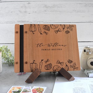 Puede incluir: Un libro de recetas de madera con una encuadernaci&oacute;n negra y un soporte. La portada est&aacute; grabada con un nombre de familia, "The Williams", y las palabras "Recetas Familiares". La portada est&aacute; decorada con una ilustraci&oacute;n en blanco y negro de varios alimentos, incluyendo verduras, pasta y hierbas.
