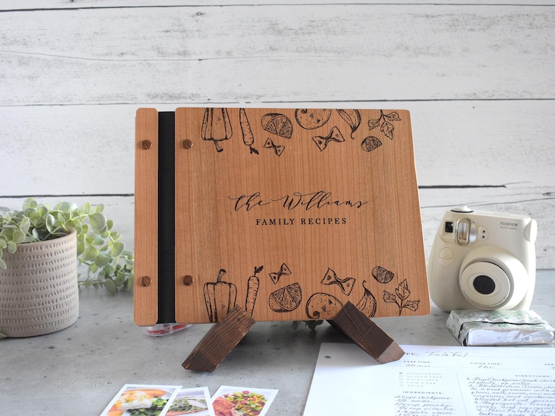 Puede incluir: Un libro de recetas de madera con una encuadernaci&oacute;n negra y un soporte. La portada est&aacute; grabada con un nombre de familia, "The Williams", y las palabras "Recetas Familiares". La portada est&aacute; decorada con una ilustraci&oacute;n en blanco y negro de varios alimentos, incluyendo verduras, pasta y hierbas.