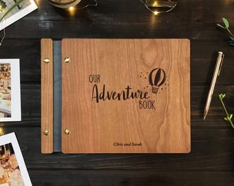 Peut inclure: Un album photo rectangulaire en bois au grain brun clair, portant le texte gravé "OUR Adventure BOOK" et une illustration de montgolfière. La couverture indique les noms "Chris and Sarah" et est reliée par des vis en métal doré.
