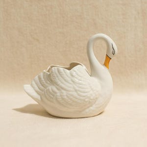May include: A white ceramic swan-shaped planter or decorative bowl featuring moulded feather details on the body, a long curved neck, and a painted orange beak with black eye markings. The piece has a smooth, glossy finish and an open top cavity.