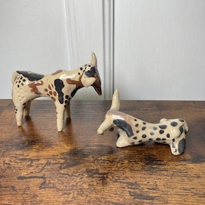 May include: Two ceramic animal figurines with a cream-coloured base and irregular black and brown spots. One figurine stands on four legs with curved horns, while the other is in a reclining position with its head turned and long, pointed horns.