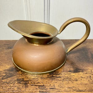 May include: A small pitcher featuring a rounded copper body and a polished brass rim, spout, and curved handle. The vessel has a wide opening and a smooth, metallic finish, showcasing a distinct contrast between the warm copper and bright brass.