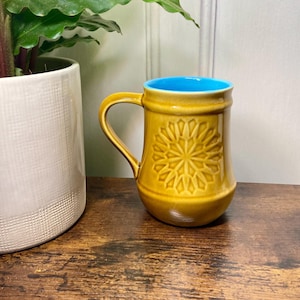 May include: Mustard yellow ceramic mug with a blue interior. The mug has a raised geometric floral design and a curved handle. The mug is on a wooden surface next to a white ceramic pot with a green plant.