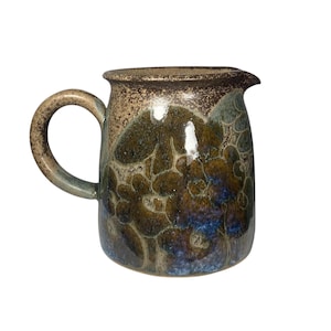 May include: A handcrafted ceramic jug with a rounded body and a curved handle. The jug features a speckled, earthy colour palette with shades of brown, green, and blue. The design has a unique, organic pattern, and the jug has a spout for easy pouring.